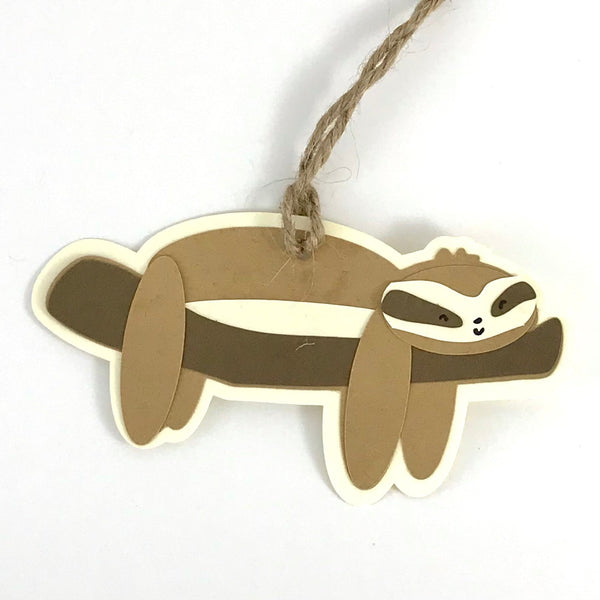 a paper gift tag in the shape of a sloth laying on top of a branch is shown against a white background