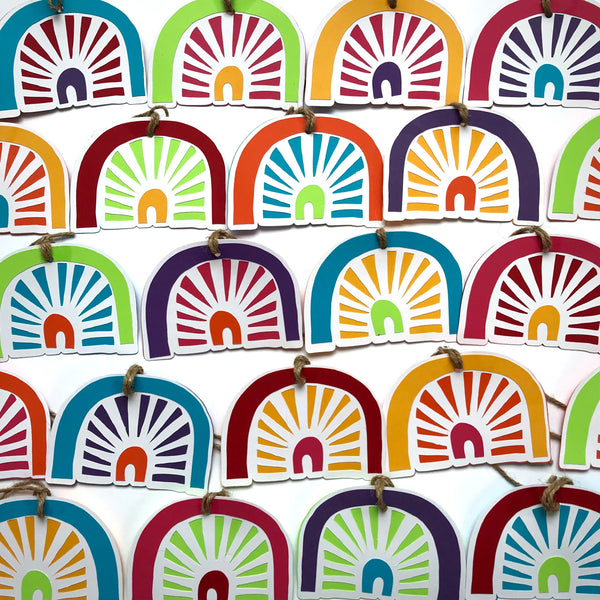 dozens of rainbow shaped paper gift tags are arranged in rows against a white background.  No two tags are the exact same colour and pattern. 