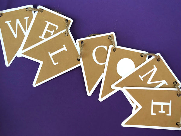 a welcome banner is shown jubbled up against a purple background.  Each letter from the banner is on it's own letter tab, cut from kraft brown paper with white paper beneather