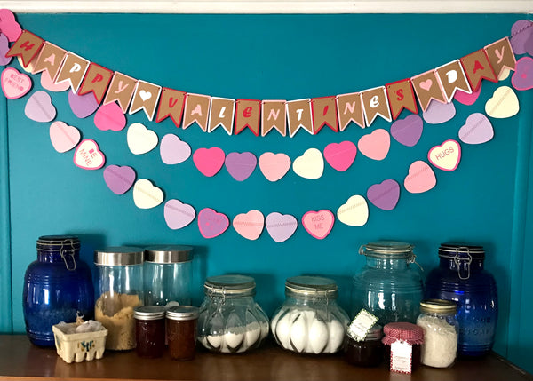 A happy valentine's day banner and two heart garlands are shown above a small pantry shelf against a blue wall.
