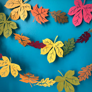 Three strings of paper leaves are hung against a blue background.  There is an assortment of oak, maple and chestnut leaves in a variety of fall colours like orange, yellow, red and more.