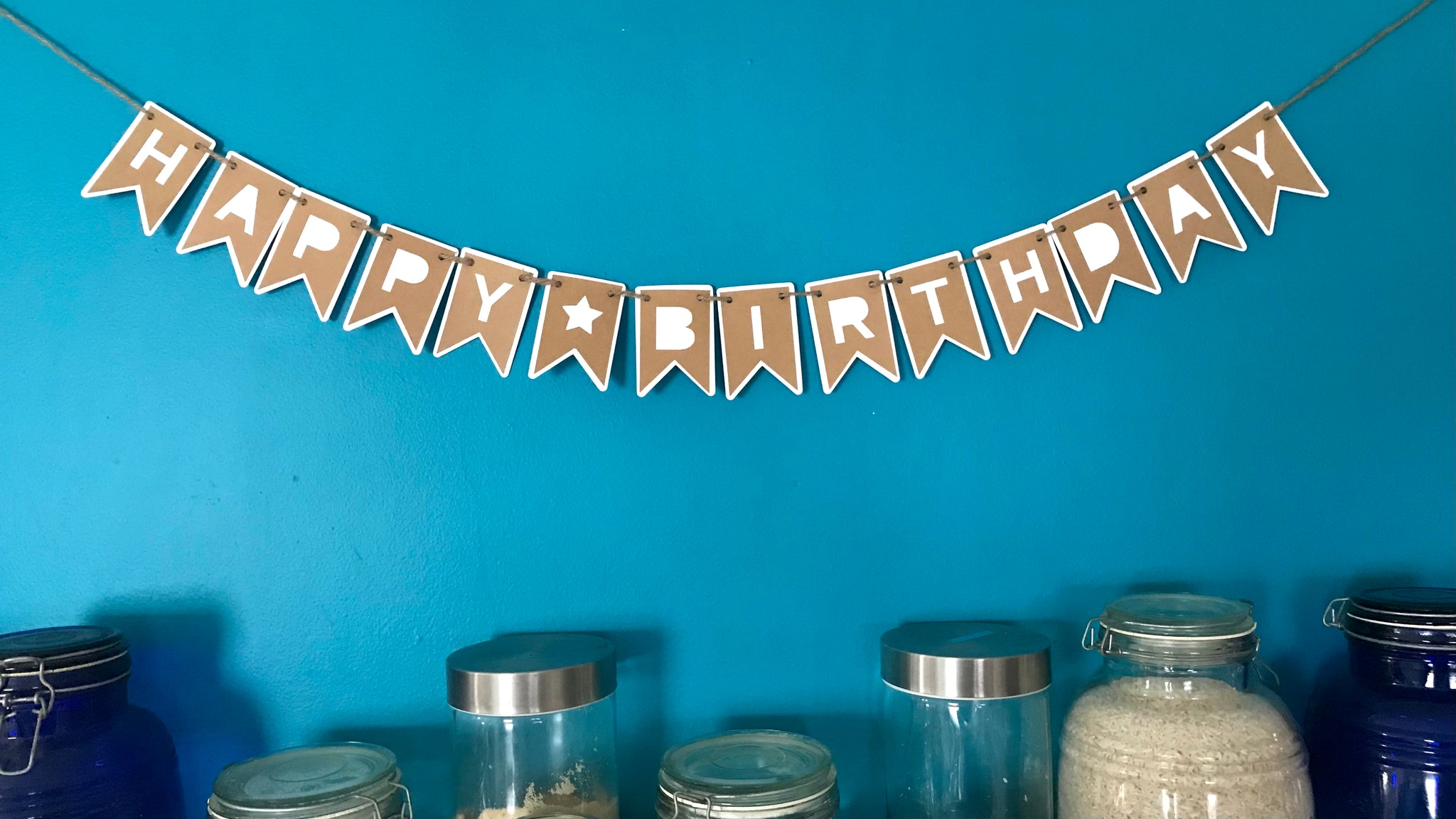 A small happy birthday banner made from kraft brown and white paper is hung against a blue wall above dry food storage.  