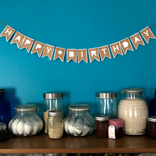 A square photo of a birthday banner hung on a blue wall above dry food storage.  Banner is made from brown and white paper.