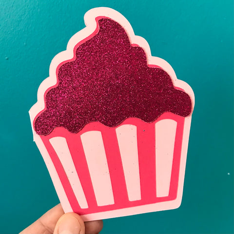 A hand holds a cupcake shaped card up against a blue background.  The cupcake is three shades of pink with glittery frosting on top