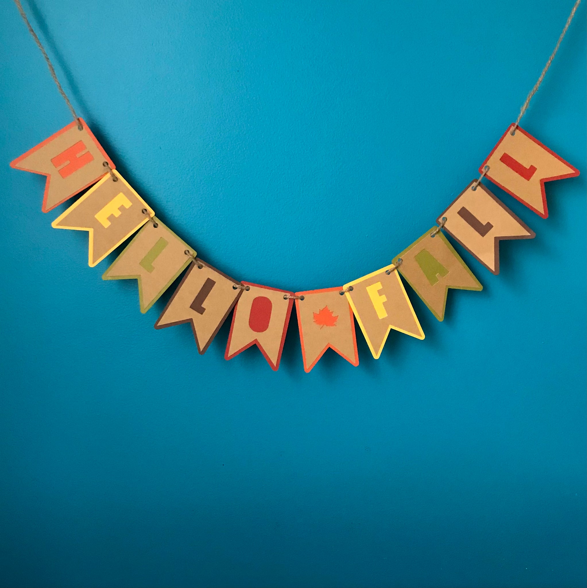 A paper banner that reads HELLO FALL is hung on hemp cord in front of a blue wall.  The Banner is cut from two layers of paper, the top layer is brown and the bottom layer is a pattern of fall colours in Orange, yellow, green, deep brown and red.