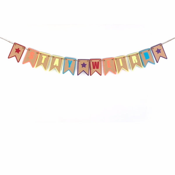 A paper banner hung on hemp cord is hanging against a white background.  The banner reads Stay Weird in a colourful rainbow pattern