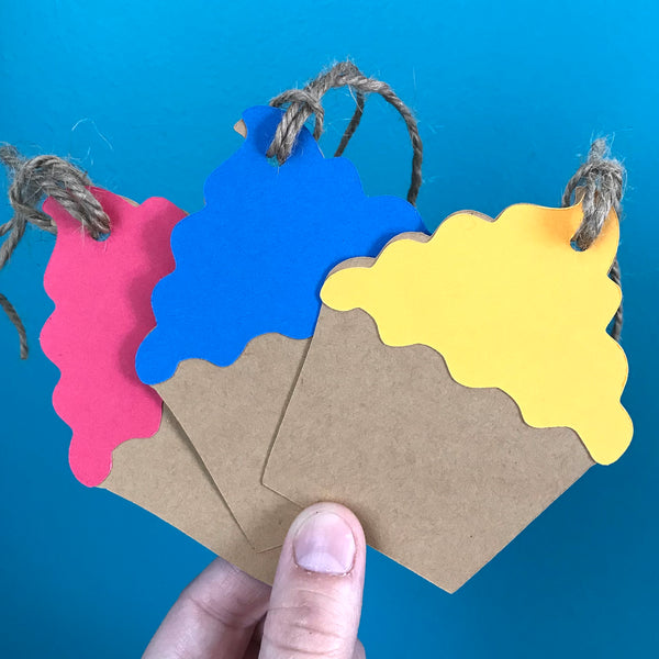 A white hand holds three cupcake shaped gift tags up against a blue background.  Each tag has hemp cord attached at the top.  The cupcakes are brown with colourful frosting, left to right frosting colours are pink, blue and yellow.