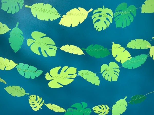 Multiple strings of green leaves are hung across a blue background.  The leaves are a variety of tropical shapes and sizes in a many different shades of green. 