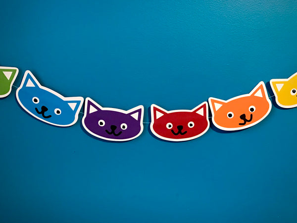 4 full kitty heads and 2 half kitty heads on a garland string are shown against a blue background.  The kitties are green, blue, purple, red, orange and yellow 