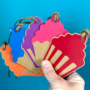 A hand holds five cupcake shaped paper gift tags with hemp cord attached up against a blue background
