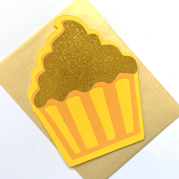 A yellow cupcake shaped card lays on top of a gold envelope and a white background. 