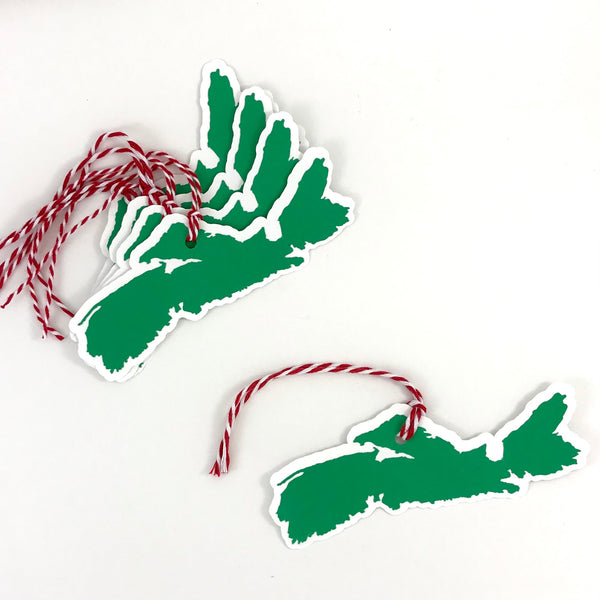 5 paper gift tags in the shape of the province of Nova Scotia are shown on a white background.  Each tag is green on white cardstock with red and white string attached.  4 tags are shown stacked together to the top left of the image with one tag shown to the bottom right of the image
