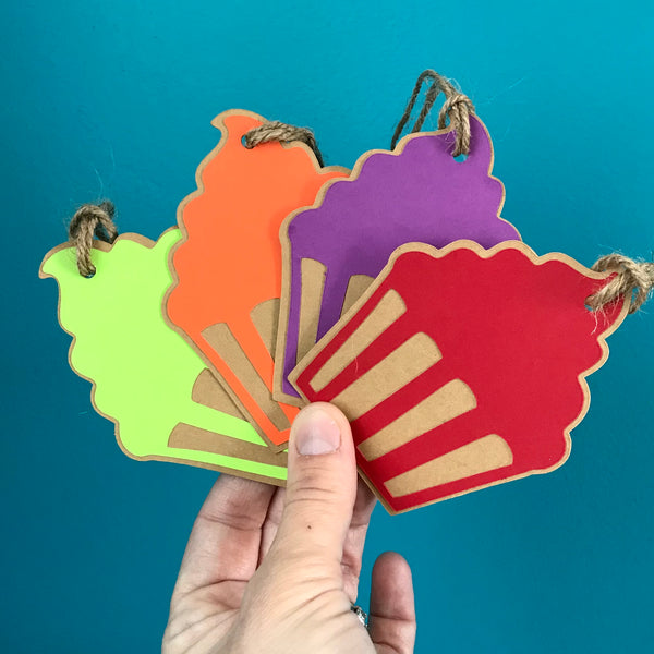 A hand holds four cupcake shaped paper gift tags up against a blue background.  Each tag has hemp cord attached at the top. The cupcakes are, left to right, green, orange, purple and red