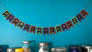 A festive banner reading Happy Fucking Halloween is hung against a blue wall above some dry food storage.  The banner is two layers of paper, the bottom layer green, purple and orange.  The top, black layer, has the letters to happy fucking halloween cut out to show the halloween colours below.