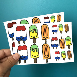 A hand holds two sheets of popsicle stickers against a blue background.  Each sheet has three different popsicle designs in three different sizes totaling 15 stickers per sheet