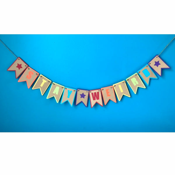 a banner that reads stay weird is hanging against a blue background.  The banner is made from two layers of paper, a brown layer and a rainbow layer, so that each letter is a different colour.  The entire banner is hung on hemp cord
