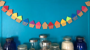 A string of 17 rainbow coloured cupcakes are hung against a blue wall above a collection of pantry storage jars.  