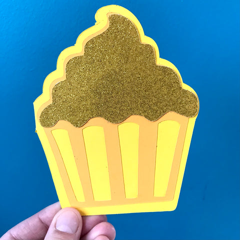 A hand holds a yellow cupcake shaped card up against a blue background.  The cupcake has glittery yellow frosting