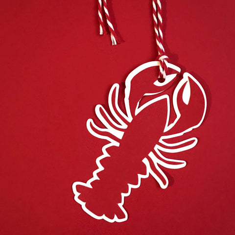 A little lobster gift tag is hanging against a red background. a red and white string is attached to the lobster's left claw.