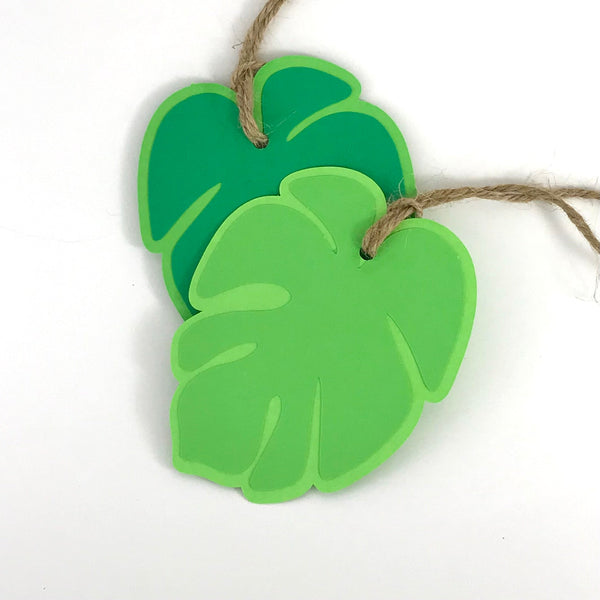 two paper gift tags in the shape of tropical leaves are shown against a white background.  each tag has hemp cord at the top