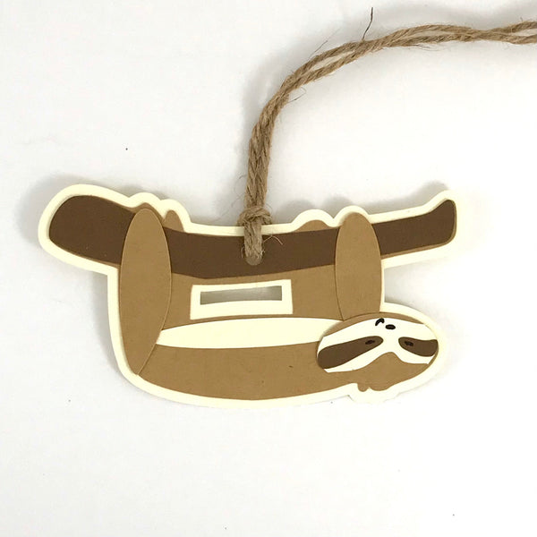 a paper gift tag in the shape of a happy little sloth is shown hanging upside down from a branch, with hemp cord at the top, against a white background
