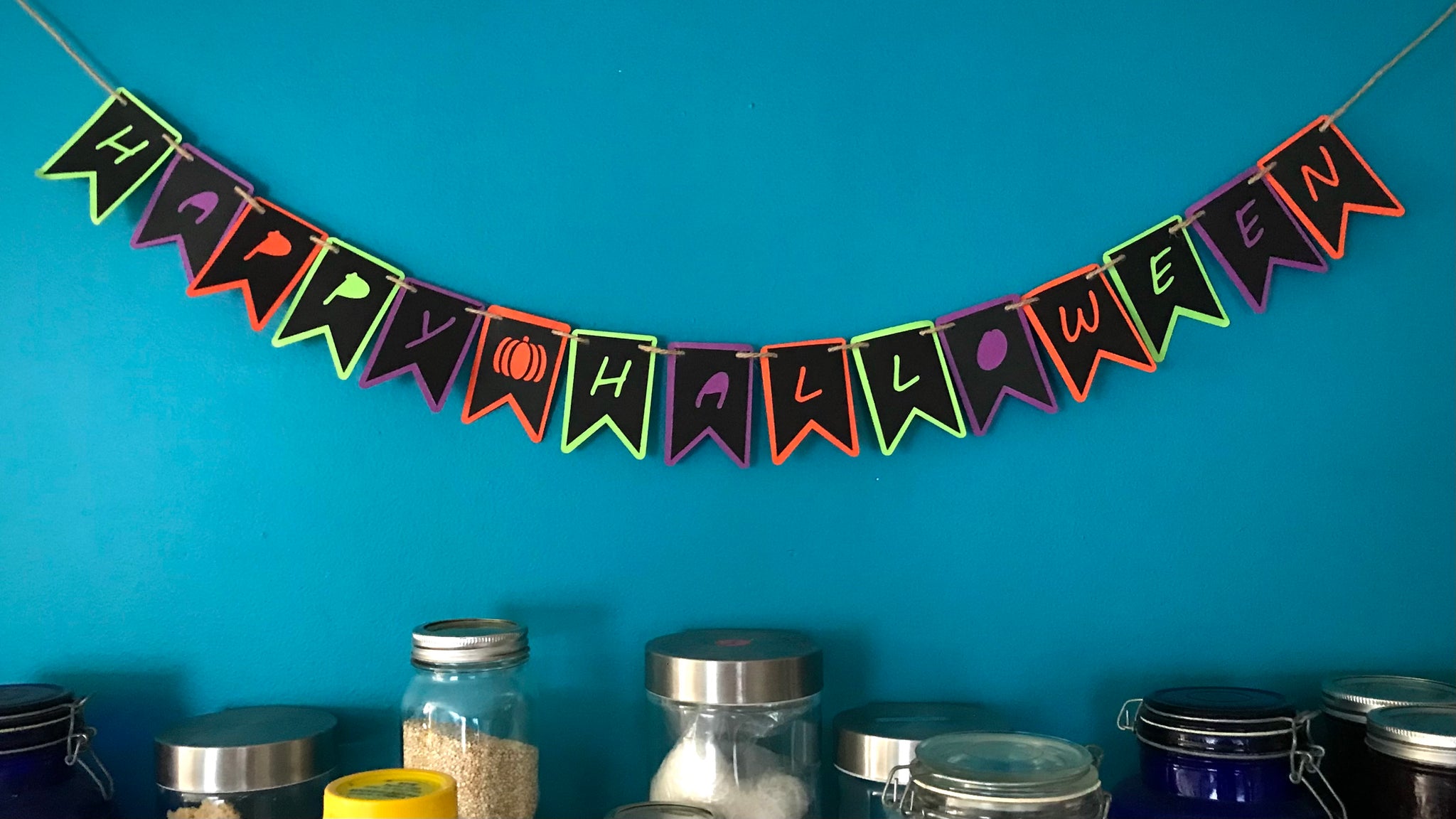 A paper banner reading Happy Halloween Is hung against a blue background over a collection of dry good storage jars.  The banner is made from two layers of paper, a top black layer with the letters cut out to show the bottom layer of green, purple and orange paper.  The banner is hung on hemp cord