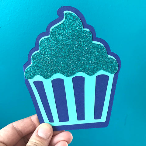 A hand holds a blue cupcake shaped card up against a blue background.  The cupcake has glittery frosting on top.