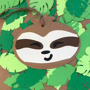 A paper gift tag in the shape of a smiling sloth face is shown against a background of tropical leaves.