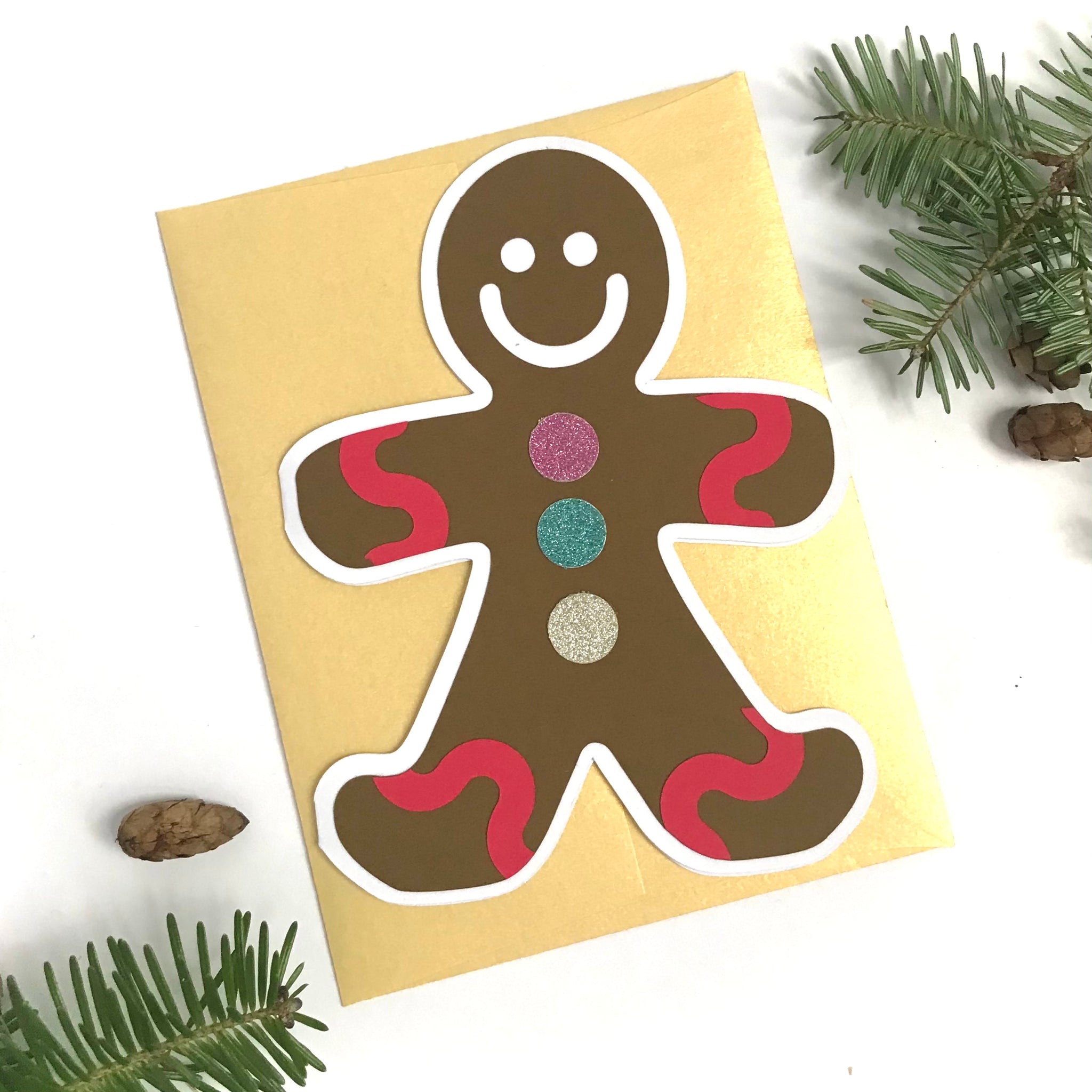 A happy little gingerbread man shaped card is laying on top of a gold envelope, against a white background with a little bit of pine tree and pine cones in the top right and bottom left corners.  The card has large glittery buttons and bright red wavy trim at the wrist and ankle of the cookie shaped card