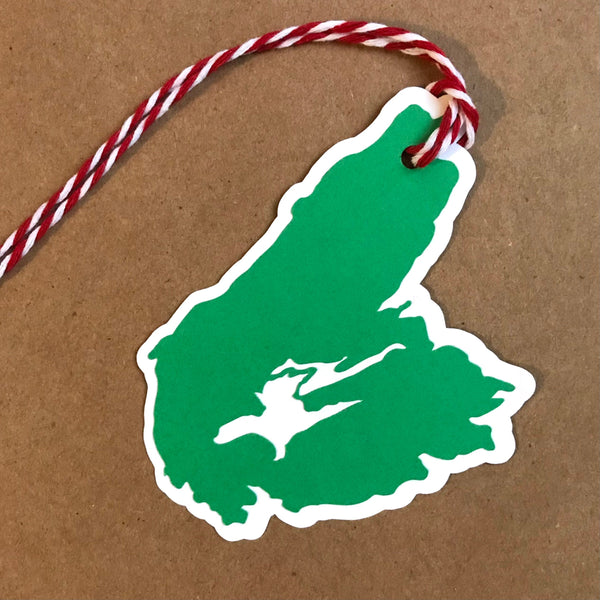 A paper gift tag in the shape of Cape Breton Island is shown against a brown background.  The tag is two layers of paper, white and green, with red and white bakers twine attached to the top of the tag.