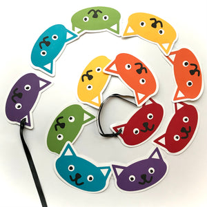 A paper garland of kitty heads is shown displayed in a swirl against a white background.  There are 12 cat heads in a repeating rainbow of colours from red to purple.