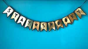 Stay Spooky! A cute banner is displayed against a blue background.  Each letter in the phrase "Stay Spooky" is cut from gold shimmery paper and mounted on black cardstock, so the letters appear black on gold.  There is a star separating the two words, and the banner is hung on black ribbon