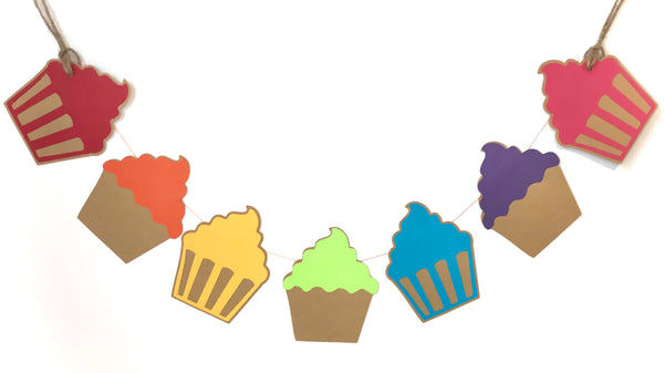 A close up of 7 cupcakes in a rainbow of colours is shown displayed against a white background