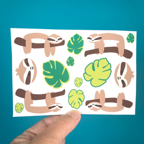 a hand holds up a single sheet of sloth and leaf stickers against a blue background