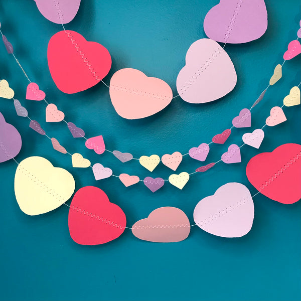 Two strings of large heart garlands and two strings of small heart garlands are shown against a blue background.  Each garland has hearts in pinks, purples and cream.
