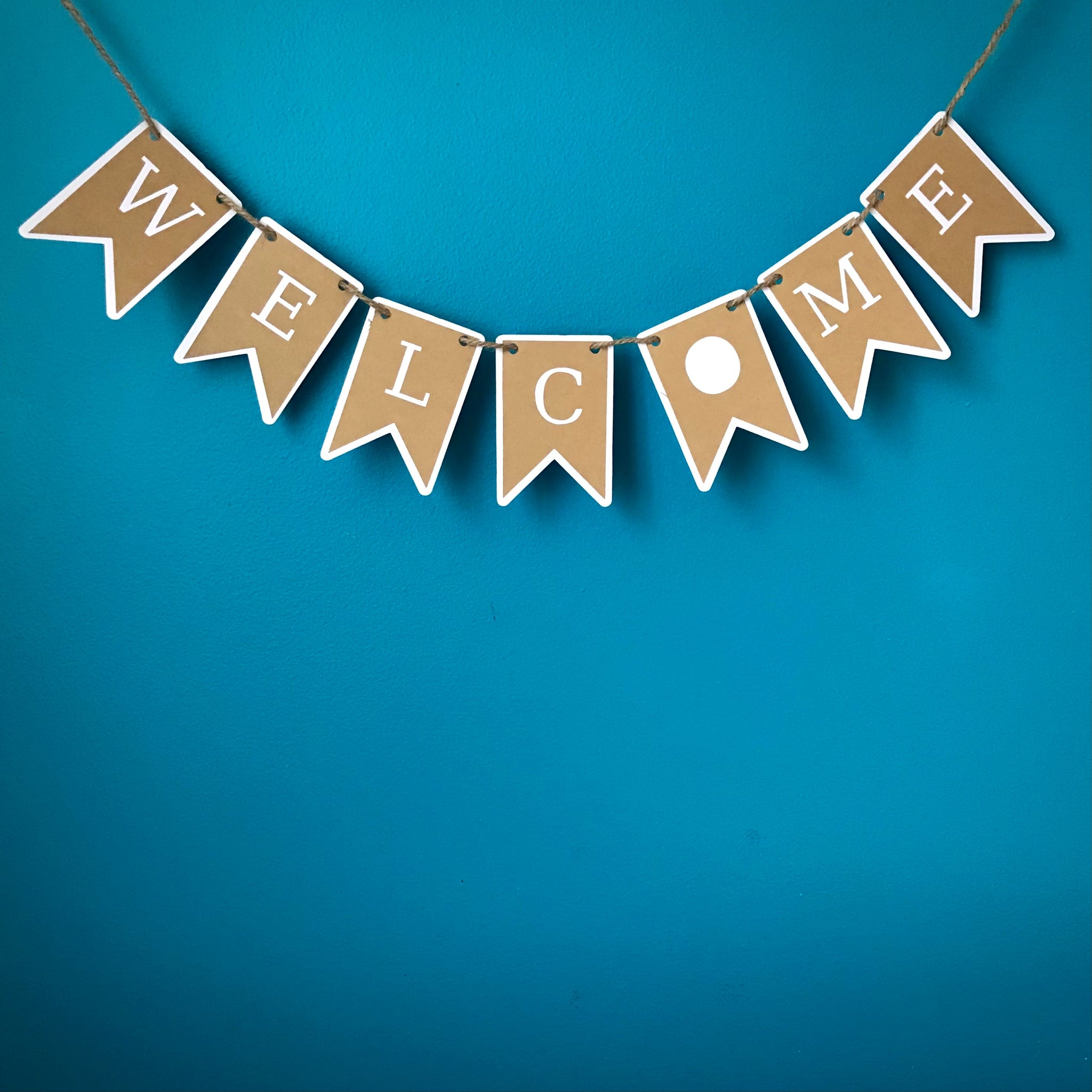A Welcome banner, shown cut from two layers of white and kraft brown paper, is shown hung on hemp cord against a blue backgroung
