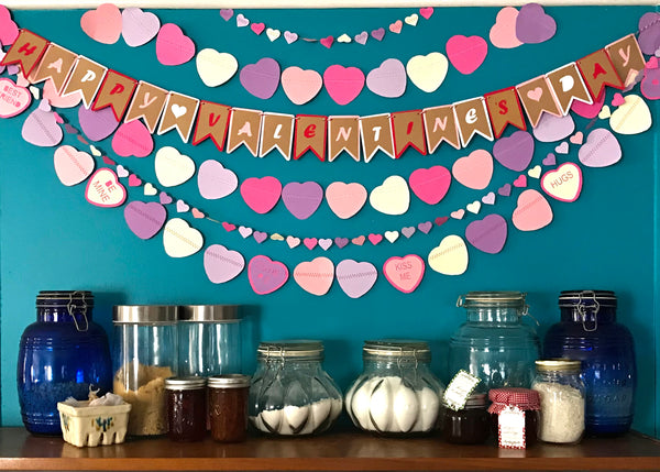 Garlands and a Happy Valentine's Day Banner are shown against a blue wall above a small dry pantry shelf.  The garlands are made from hearts in a variety of sizes, all in pinks and purples.